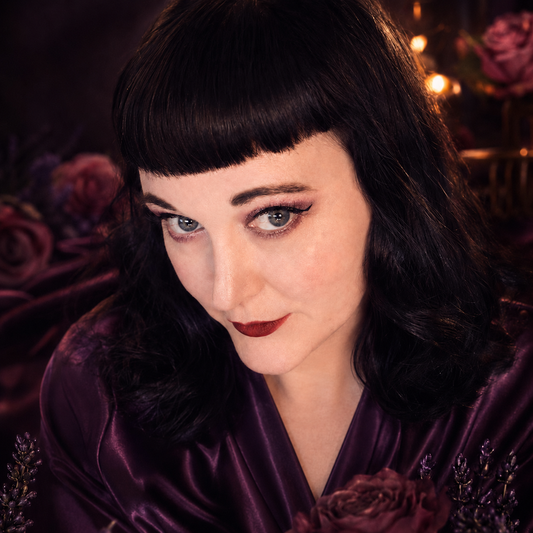 Woman in a purple satin robe with dramatic eye makeup, surrounded by dark roses and warm candlelight.