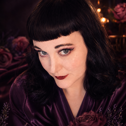 Woman in a purple satin robe with dramatic eye makeup, surrounded by dark roses and warm candlelight.