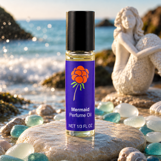 Close-up of Cloudberry Beauty Mermaid Perfume Oil displayed by the sea with sea glass, shells and a mermaid statue.