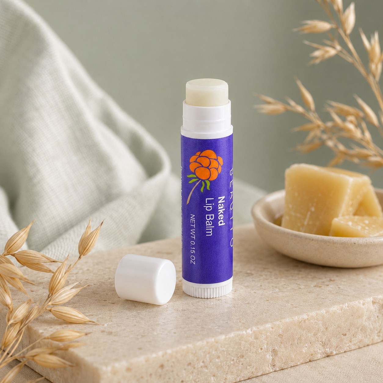 Cloudberry Beauty Naked Lip Balm tube displayed beside beeswax and dried oat stems on a stone surface.